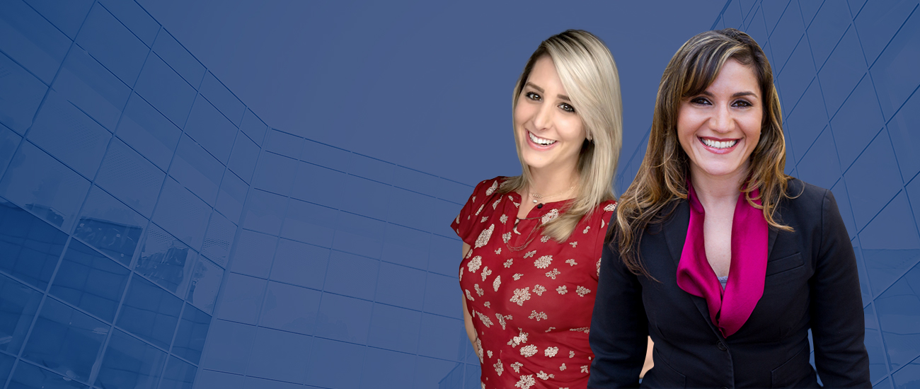 Photo of Rina Feld and Hailey Wilson standing in front of a modern building with a reflective glass facade under a clear sky.